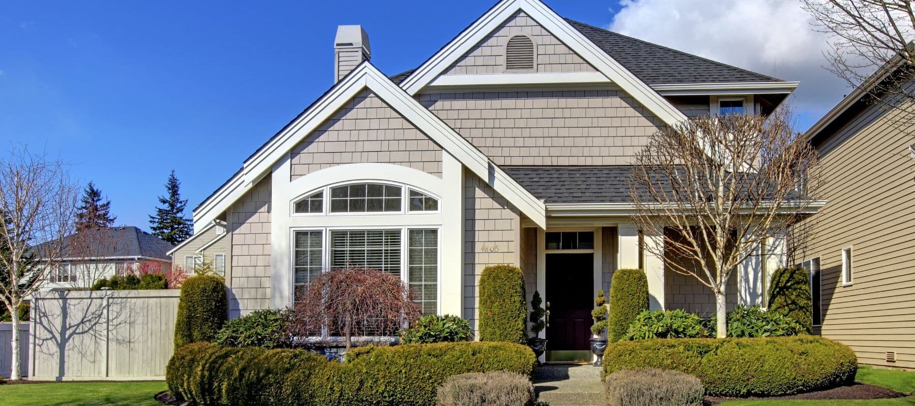 Light brown shingle-style house with manicured shrubs and arched front window